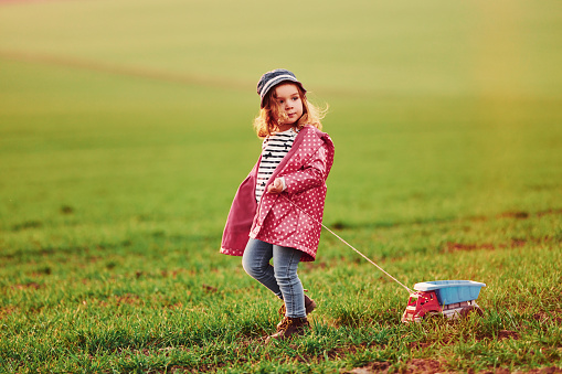 Cute little girl walks with toy car on the beautiful field at sunny daytime.
