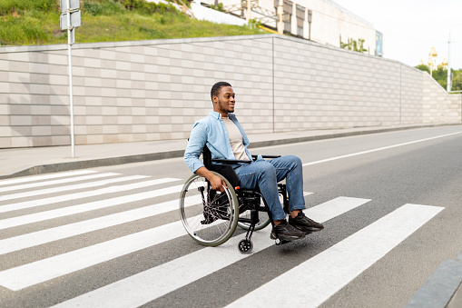 Retrato de cuerpo entero de un joven negro discapacitado en silla de ruedas cruzando la calle de la ciudad, espacio de copia photo