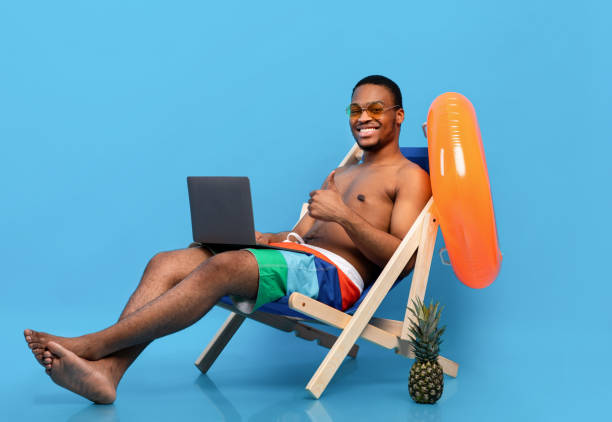 istock Cheerful black guy using laptop, browsing internet during beach vacation, sitting in lounge chair, showing thumb up 1326413317