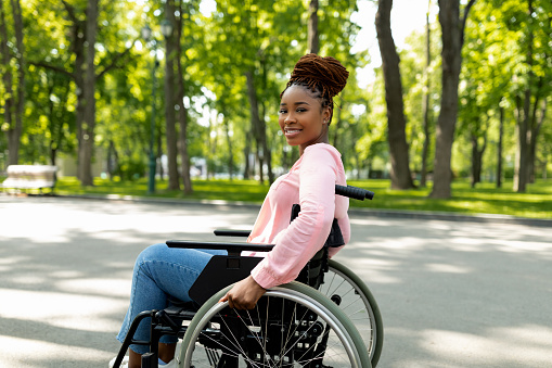 Positive handicapped black lady in wheelchair enjoying wonderful summer morning outdoors. Happy disabled African American woman having great time at city park. Urban lifestyle