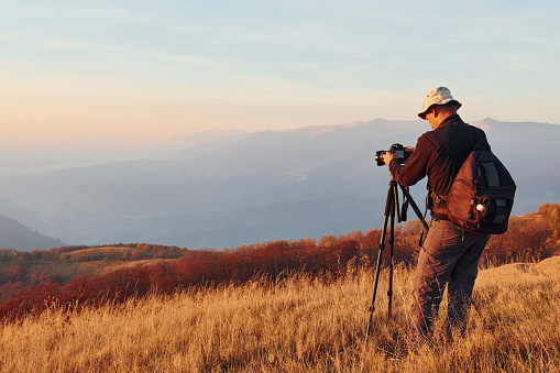 Male photographer standing and working at majestic landscape of autumn trees and mountains by the horizon.