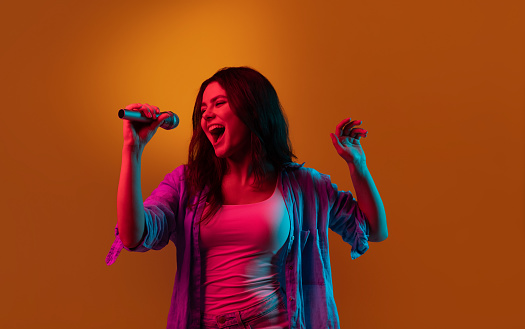 Caucasian female singer portrait isolated on blue studio background in neon light. Beautiful female model with microphone. Concept of human emotions, facial expression, ad, music, art.