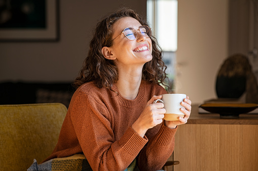 Portrait of joyful young woman enjoying a cup of coffee at home. Smiling pretty girl drinking hot tea in front of the window in winter. Excited woman wearing spectacles and sweater and laughing in an autumn day.