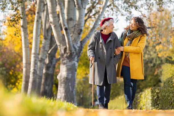 Woman with grandmother walking in park in autumn Granddaughter walking with senior woman in park wearing winter clothing. Old grandmother with walking cane walking with lovely caregiver girl in sunny day. Happy woman and smiling grandma walking in autumn park. adult age stock pictures, royalty-free photos & images