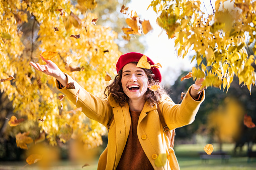 Beautiful young woman throwing autumn yellow leaves. Portrait of young woman playing with leaves in park during fall. Beautiful girl wearing coat and red beret playing with yellow leaves outdoor during foliage season.