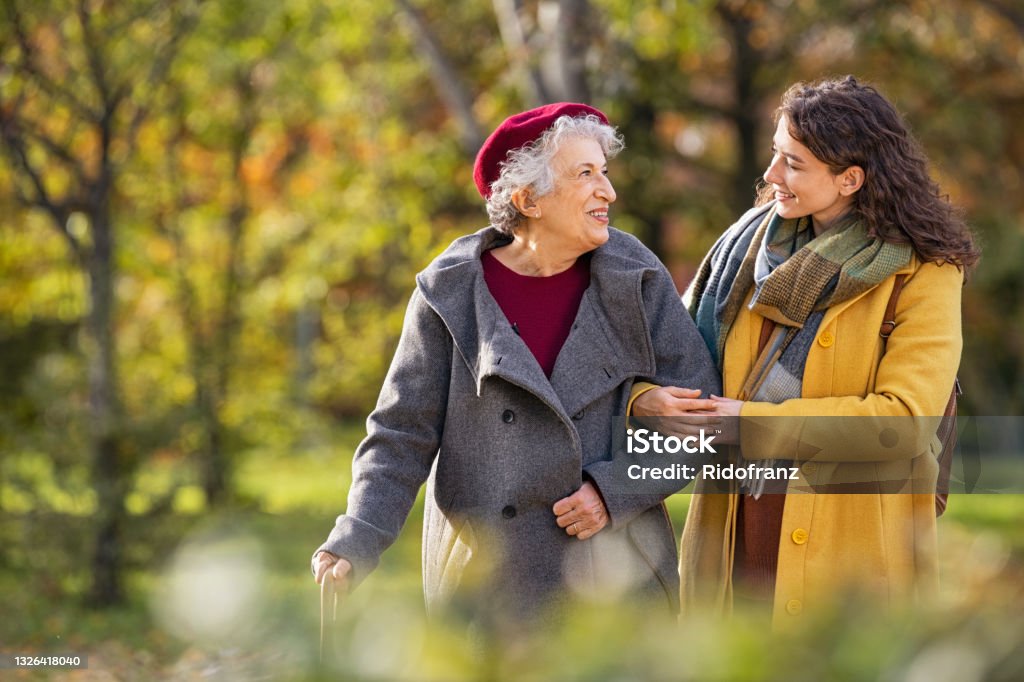 Senior woman walking with granddaughter in park during autumn Young woman in park wearing winter clothing walking with old grandmother. Happy grandma wearing coat walking with lovely girl outdoor with copy space. Smiling lovely caregiver and senior lady walking in park during autumn and looking at each other. Senior Adult Stock Photo