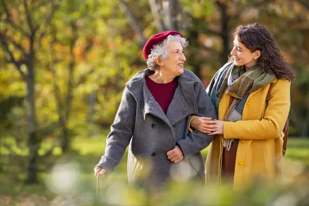 Young woman in park wearing winter clothing walking with old grandmother. Happy grandma wearing coat walking with lovely girl outdoor with copy space. Smiling lovely caregiver and senior lady walking in park during autumn and looking at each other.