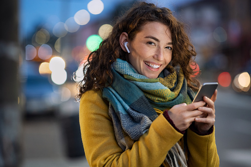 Young beautiful woman using her mobile phone at dusk in the city while listening music through earphones. Smiling girl sending message from cellphone in urban environment. Happy woman using smartphone to do a video call on a winter day while looking at camera.