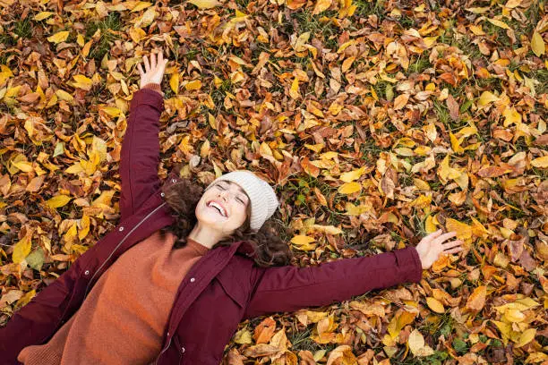High angle view of young playful woman lying over autumn leaves with stretched arms and copy space. Happy beautiful girl wearing jacket lying over dry leaves and smiling with closed eyes. Young woman rest on autumn foliage in park and having fun.