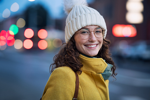 Portrait of happy young woman wearing warm hat with eyeglasses on city street and looking at camera. Pretty girl standing outdoors on winter evening with a toothy smile and copy space. Beautiful student with cap and winter coat walking at urban road at dusk.