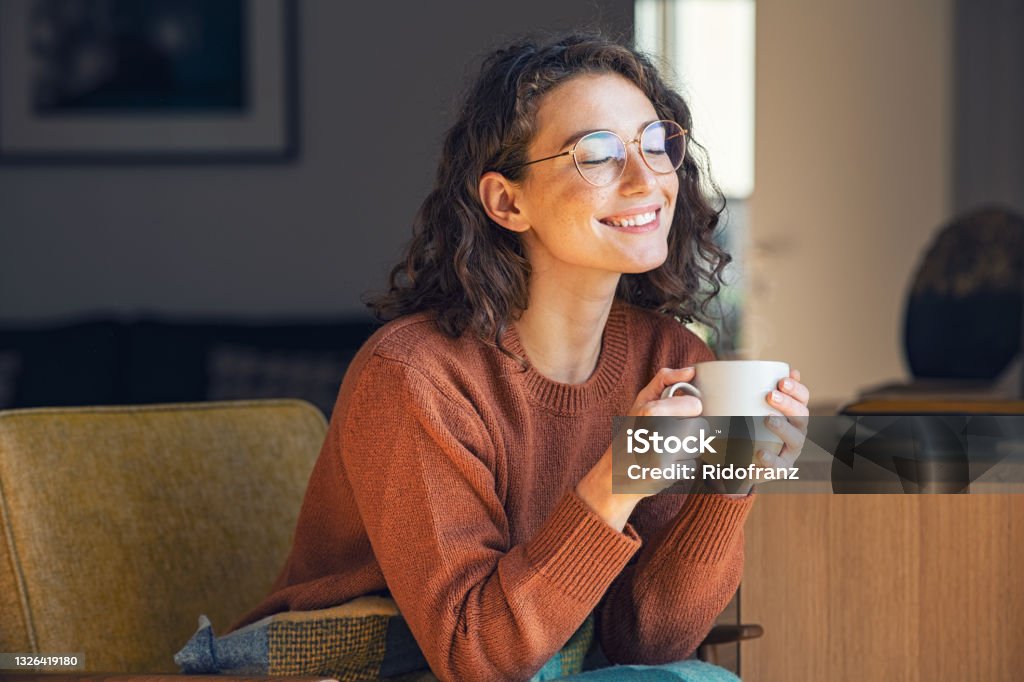 Beautiful woman relaxing and drinking hot tea Happy young woman drinking a cup of tea in an autumn morning. Dreaming girl sitting in living room with cup of hot coffee enjoying under blanket with closed eyes. Pretty woman wearing sweater at home and enjoy a ray of sunshine on a winter afternoon. Coffee - Drink Stock Photo