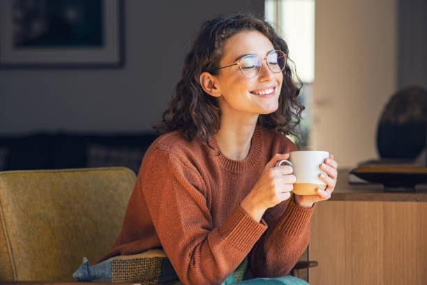 Beautiful woman relaxing and drinking hot tea Happy young woman drinking a cup of tea in an autumn morning. Dreaming girl sitting in living room with cup of hot coffee enjoying under blanket with closed eyes. Pretty woman wearing sweater at home and enjoy a ray of sunshine on a winter afternoon. cup stock pictures, royalty-free photos & images