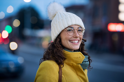 Smiling young woman wearing warm hat with eyeglasses on city street waiting for the bus. Happy girl wearing yellow coat and standing outdoors on winter evening at bus stop. Carefree happy girl in cold urban center during dusk.