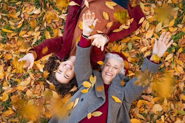 Grandmother and granddaughter lying on foliage and enjoy the autumn Senior grandmother and smiling granddaughter lying on yellow and red leaves at park, view from above. Top view of playful old woman with cheerful girl lying on autumn park leaves. High angle view of happy grandmother and young woman playing with dried leaves while lying on ground at park and looking at camera. throwing photos stock pictures, royalty-free photos & images