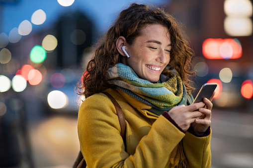 Young beautiful woman using her mobile phone while listening voice note with wireless earbuds. Smiling girl changing songs from smart phone in urban environment. Happy smiling woman chatting on smartphone on a winter day in urban road.