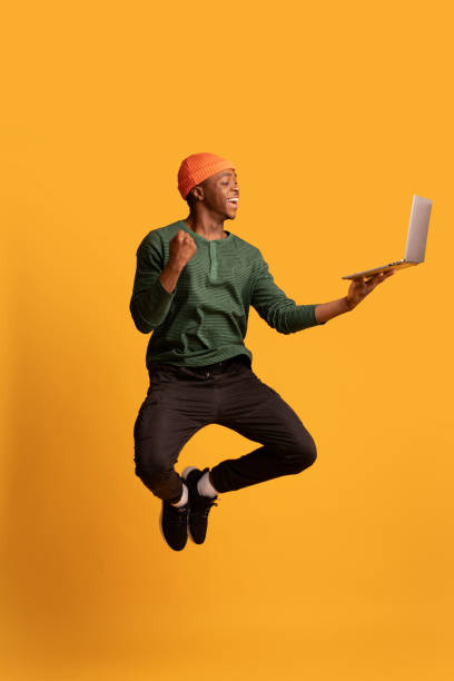 Online Win. Excited Happy Black Guy Jumping Up With Laptop Computer Online Win. Excited Happy Black Guy Jumping Up With Laptop Computer, Emotional Young African American Man Celebrating Success Over Yellow Studio Background, Full Length Shot With Free Space hipster person stock pictures, royalty-free photos & images