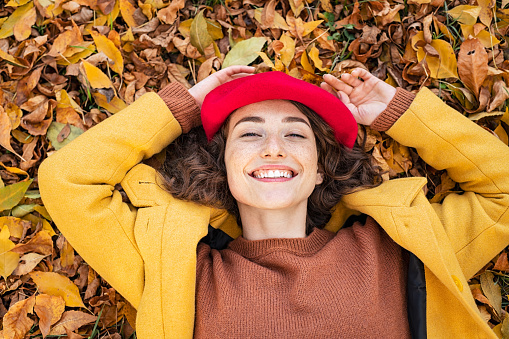 Happy smiling woman lying over yellow leaves and looking at camera. Young beautiful woman lying on dry autumn leaves with a big grin. Portrait of cheerful girl rest on ground in park with hands behind head.