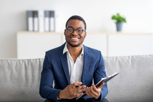 Portrait of happy black male psychologist looking at camera and taking notes during therapy session at clinic Portrait of happy black male psychologist looking at camera and taking notes during therapy session at clinic. Friendly millennial African American psychotherapist posing and smiling at office mental health professional stock pictures, royalty-free photos & images