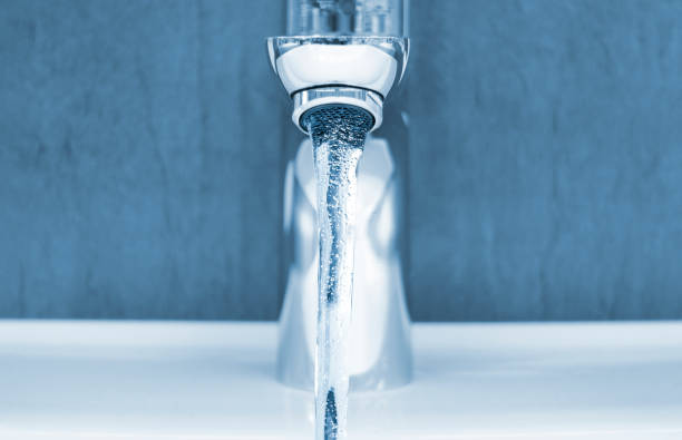 Front view of a modern open tap with running water in bathroom at home. \nIn the foreground the detailed water jet. in the background copy space. blue toned image