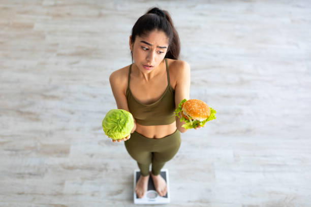 Above view of young Indian woman holding cabbage and hamburger, standing on scales, making choice of her meal