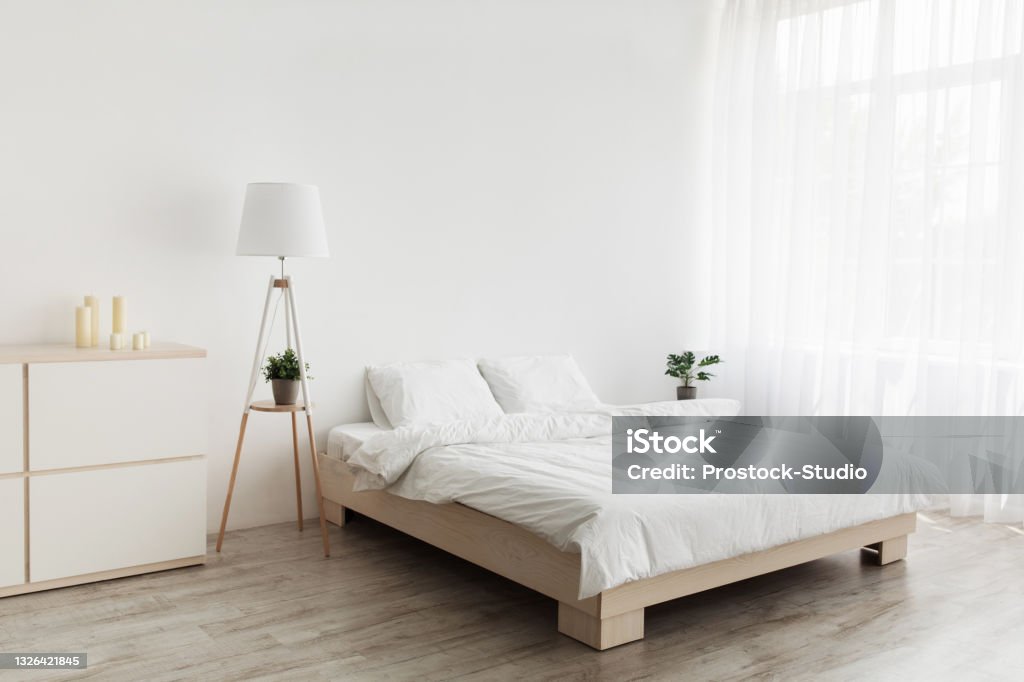 Simple modern design, ad, offer. Double bed with white pillows and soft blanket, lamp, furniture on wooden floor Simple modern design, ad, offer. Double bed with white pillows and soft blanket, lamp, furniture on wooden floor. White empty wall, big window with curtains in bedroom interior Sparse Stock Photo