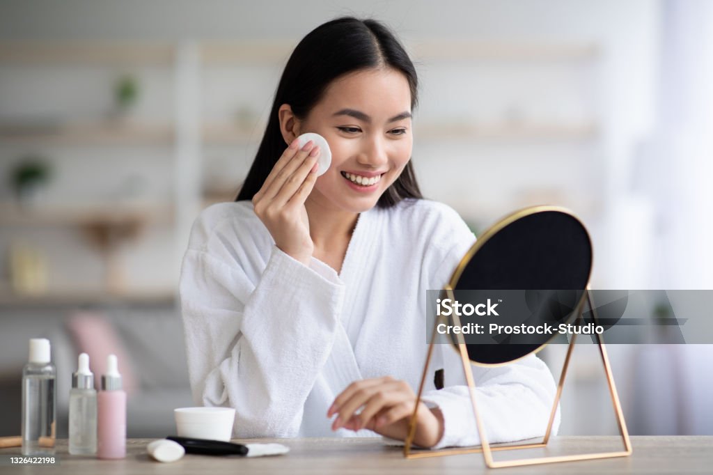 Beautiful asian woman cleaning her face with toner Beautiful asian woman cleaning her face, using cotton pads and cleansing product, looking at mirror in bedroom. Young attractive korean lady using face toner and cotton pad, home interior, empty space Skin Care Stock Photo