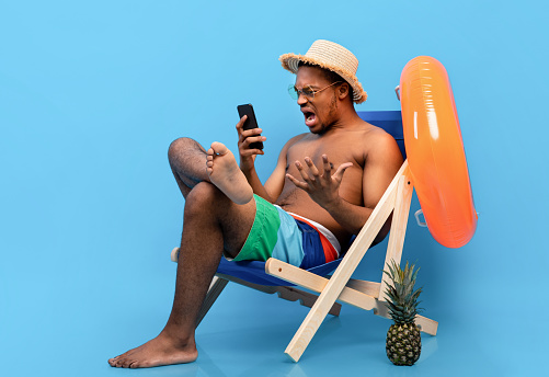 Frustrated black guy shouting at smartphone, feeling stressed, having bad internet connection on summer vacation, sitting in lounge chair with mobile device, blue studio background