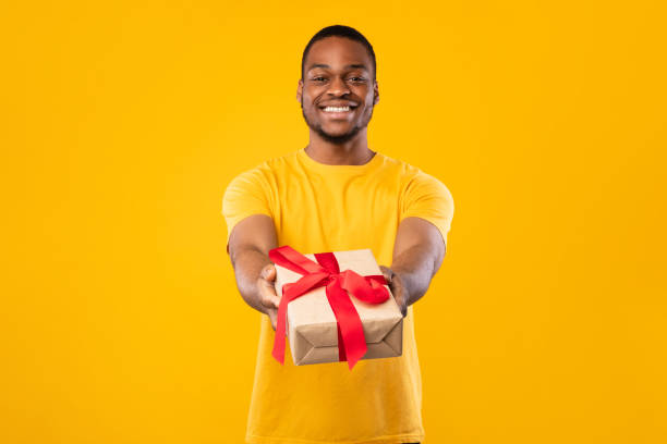 istock Black Guy Giving Wrapped Present Box To Camera, Yellow Background 1326422366