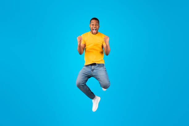 Emotional Black Guy Shouting Gesturing Yes In Mid-Air, Blue Background Joy Of Victory. Emotional Black Guy Gesturing Yes And Shouting Posing In Mid-Air Over Blue Background. Millennial Man Jumping And Shaking Fists Looking At Camera Celebrating Success In Studio jumping stock pictures, royalty-free photos & images