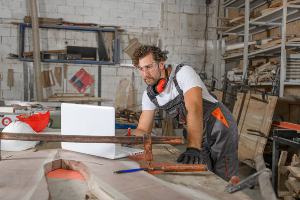 A Male Carpenter in Work Uniform and Protective Workwear is Doing Manufacturing Job in Large Workshop While Using a Laptop for Online Help.