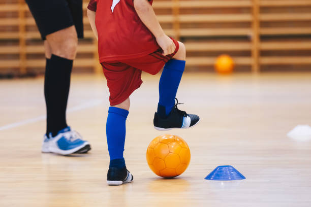 オーナジフットサルボールとサッカー服を着た少年。コーチと屋内サッカーのトレーニングで子供。木の床でフットサルをする子供。コーチトレーナーとのフットサルスクールの練習 - futsal indoors soccer ball soccer ストックフォトと画像