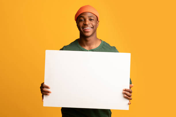 allegro ragazzo nero che tiene un cartello bianco bianco mentre posa su sfondo giallo - holding a sign foto e immagini stock