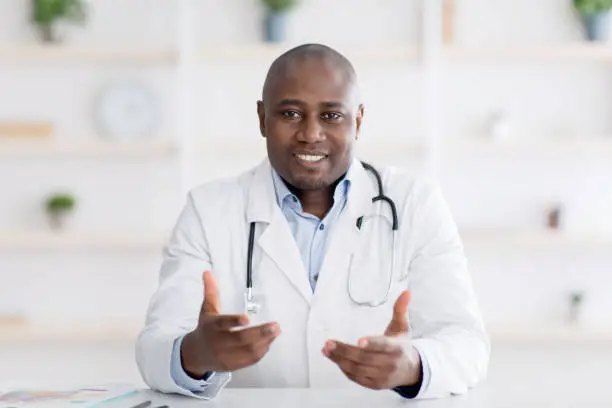 African american doctor sitting at workdesk at clinic, having appointments during coronavirus pandemic, giving recommendations. Male doc sitting at cabinet in hospital, copy space