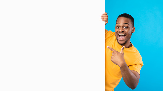 Great Sales Offer. Excited African American Man Pointing Finger At Copy Space On Big White Paper Poster Advertising Your Text Over Blue Studio Background, Smiling To Camera. Panorama, Mockup