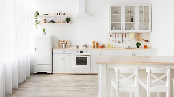 Family dining room and minimalist kitchen interior. Wooden table, white chairs, kitchenware and utensils on furniture and shelves, small refrigerator, range hood, white walls, panorama