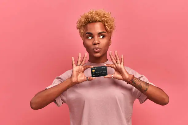 Photo of Portrait of a beautiful young African American woman with blond curly hair wearing lilac clothes holding a credit card with clipping path, looks away, thinking about purchases, isolated over pink wall