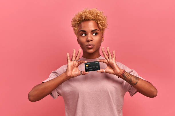 portrait of a beautiful young african american woman with blond curly hair wearing lilac clothes holding a credit card with clipping path, looks away, thinking about purchases, isolated over pink wall - cartão de saudações imagens e fotografias de stock