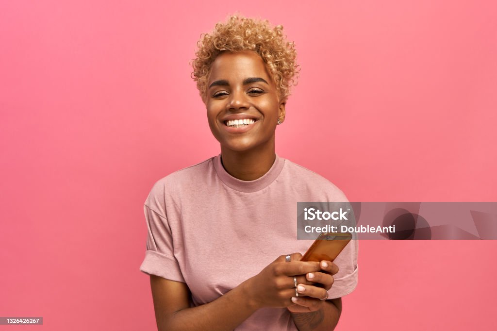Halblange Aufnahme von positiven attraktiven weiblichen Modell mit Afro-Haarschnitt, fühlt sich gut, nutzt Smartphone-Gerät für Unterhaltung und Online-Chat, Surfer Social-Network-Profil, nutzt kostenloses Internet. - Lizenzfrei Frauen Stock-Foto