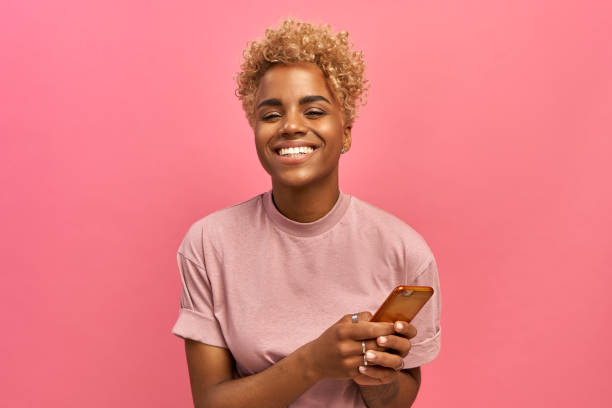 halblange aufnahme von positiven attraktiven weiblichen modell mit afro-haarschnitt, fühlt sich gut, nutzt smartphone-gerät für unterhaltung und online-chat, surfer social-network-profil, nutzt kostenloses internet. - lächeln stock-fotos und bilder