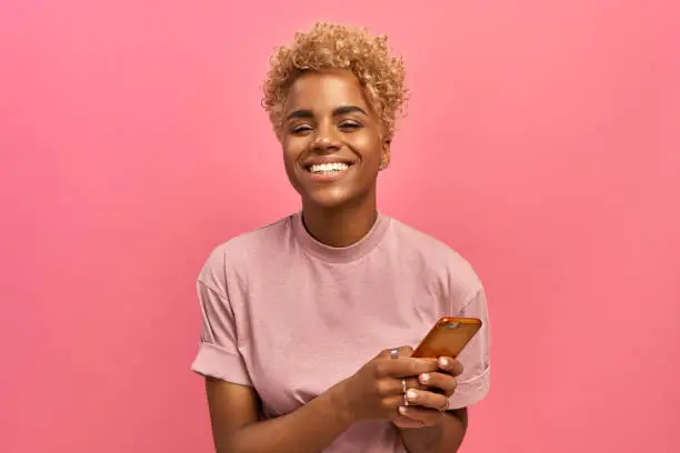 Photo of Half length shot of positive attractive female model with Afro haircut, feels good, uses smartphone device for entertainment and online chatting, surfers social network profile, uses free internet.