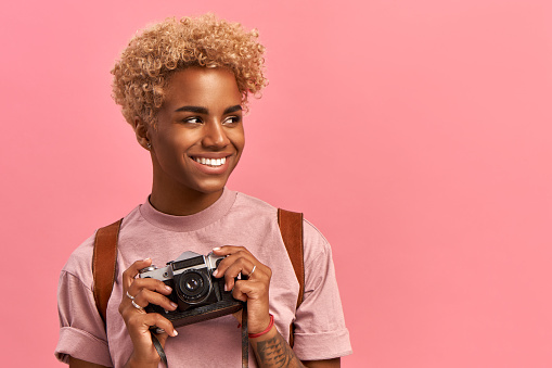 Image of talented Afro female photographer makes professional shots during adventure trip, uses retro camera, wears lavender t-shirt and backpack, enjoys holidays, poses indoor. Active rest concept.