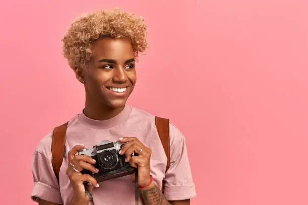Photo of Image of talented Afro female photographer makes professional shots during adventure trip, uses retro camera, wears lavender t-shirt and backpack, enjoys holidays, poses indoor. Active rest concept