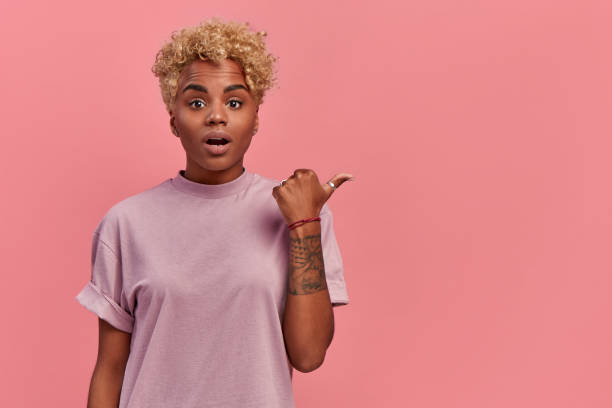 istock Attractive young African American female points aside with surprised expression, feeling astonished excited advertising product or service isolated on pink background. Advertisement and ethnicity. 1326426293