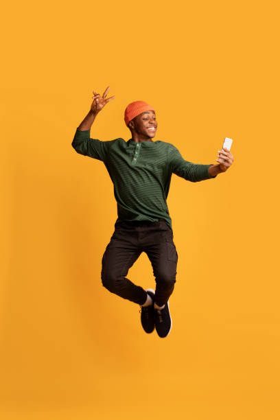 공중에서 점프하는 동안 스마트 폰으로 셀카를 찍는 쾌활한 아프리카 계 미국인 남자 - photography vertical color image studio shot 뉴스 사진 이미지