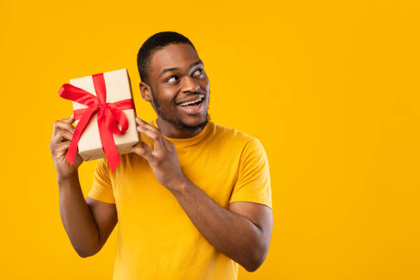 istock Funny African Guy Holding Gift Guessing What's Inside, Yellow Background 1326427456