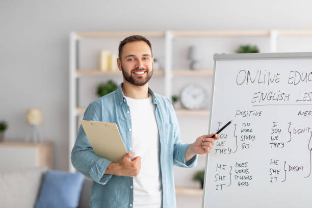 joyeux jeune enseignant masculin pointant vers le tableau noir, expliquant la grammaire, donnant une leçon d’anglais en ligne depuis le bureau à domicile - langue anglaise photos et images de collection