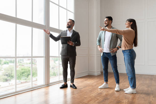 Estate Agent Showing Prospective Buyers Around Property Professional Real Estate Agent Showing Spacious New Apartment To Young Couple, Looking And Pointing At Beautiful View From Big Panoramic Picture Windows. Happy Man And Woman Ready to Become Homeowners visit stock pictures, royalty-free photos & images