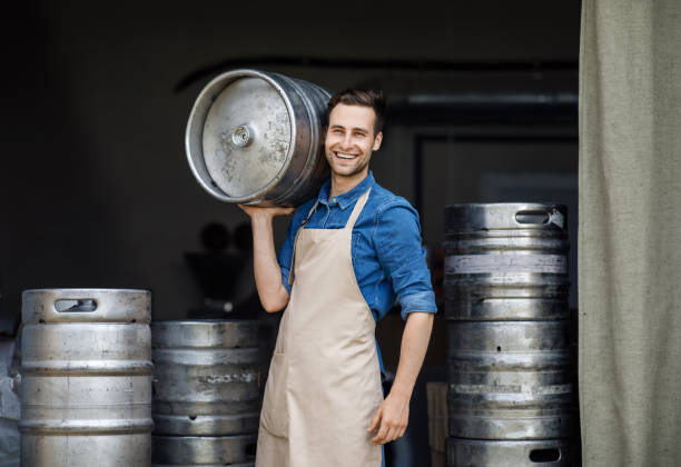 Attractive confident strong man working at plant. Owner of brewery, employee at storage Attractive confident strong man working at plant. Owner of brewery, employee at storage. Happy young attractive guy in apron carries large barrel in warehouse with metal kegs, on black background man wearing a barrel stock pictures, royalty-free photos & images