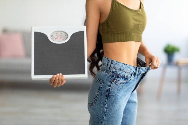 cropped view of indian woman in oversized jeans holding scales, impressed with results of weight loss diet - east asian ethnicity imagens e fotografias de stock