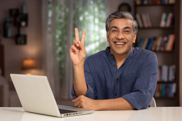 shot of a mature business men working at home using laptop:- stock photo Adult, adult only, business person, mature men India, Indian ethnicity, peace sign gesture photos stock pictures, royalty-free photos & images